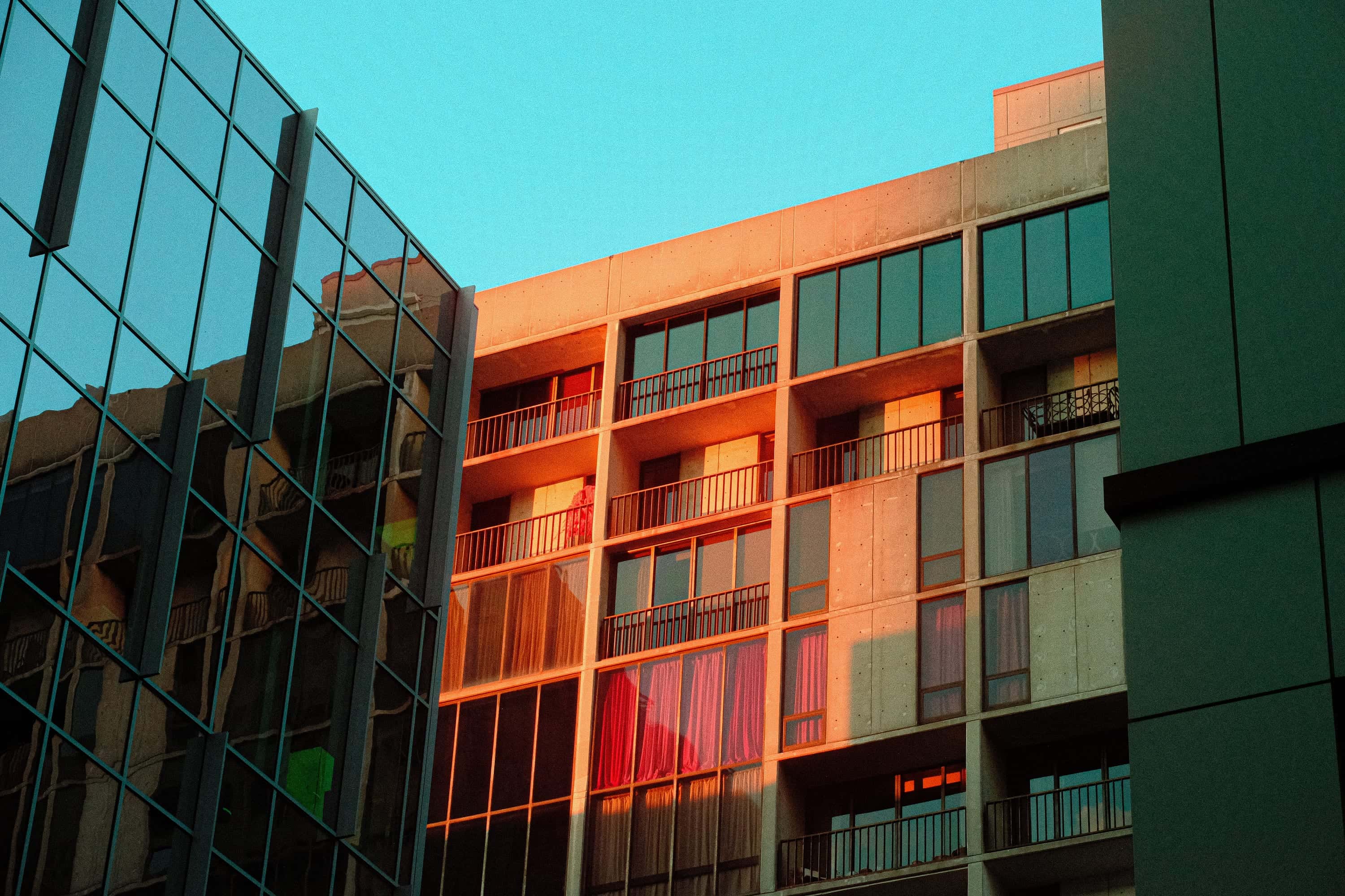 Two facades at sunset, one glass reflecting teal, the other concrete balconies lit molten orange