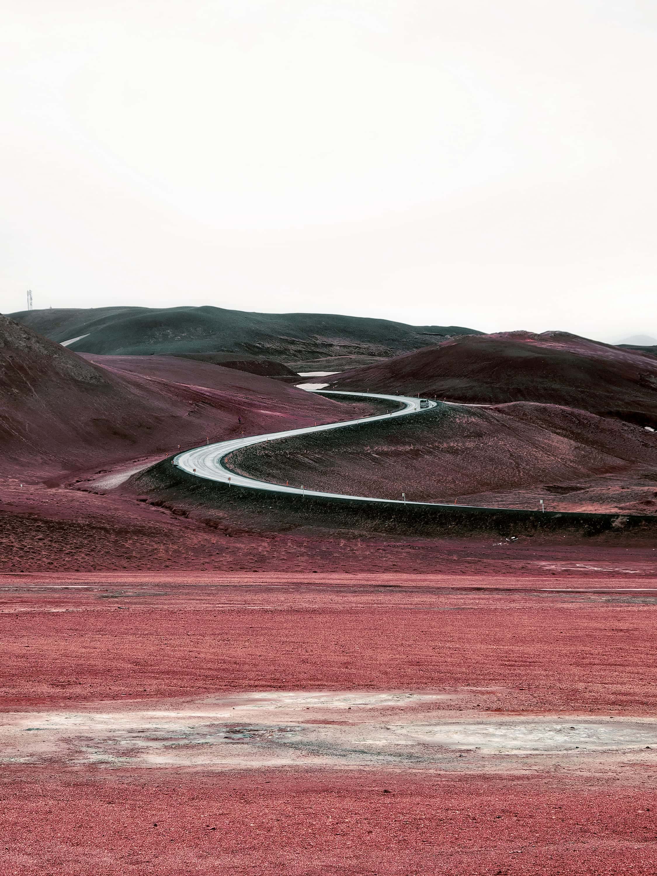 A paved road curving in a long S through bare rounded hills, with a single car on it and a mast on the ridge behind, photographed in near-infrared light.
