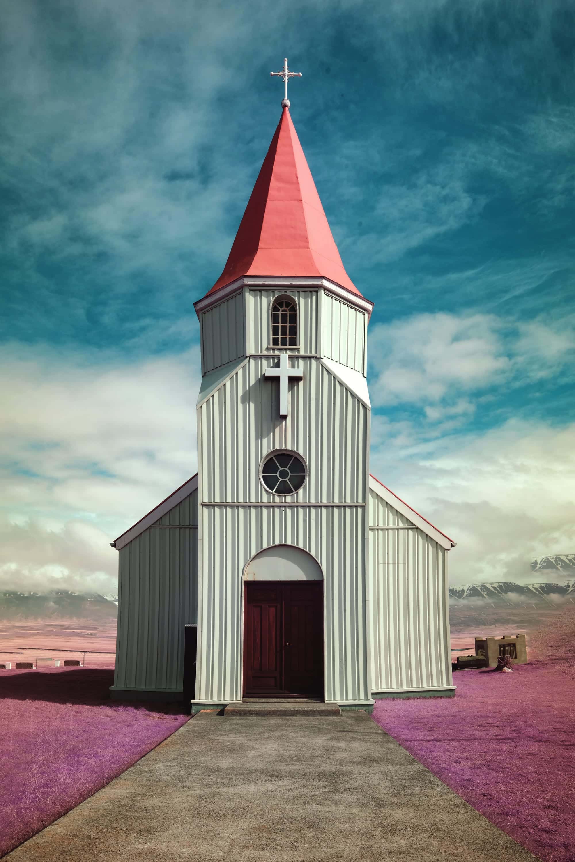 The church at Glaumbær in Skagafjörður, northern Iceland, its corrugated-iron walls and tall spire standing alone on the valley floor below snow-streaked mountains, photographed in near-infrared light. The churchyard grass carries the strongest signal in the frame while the metal cladding and the gravel path sit flat beside it.