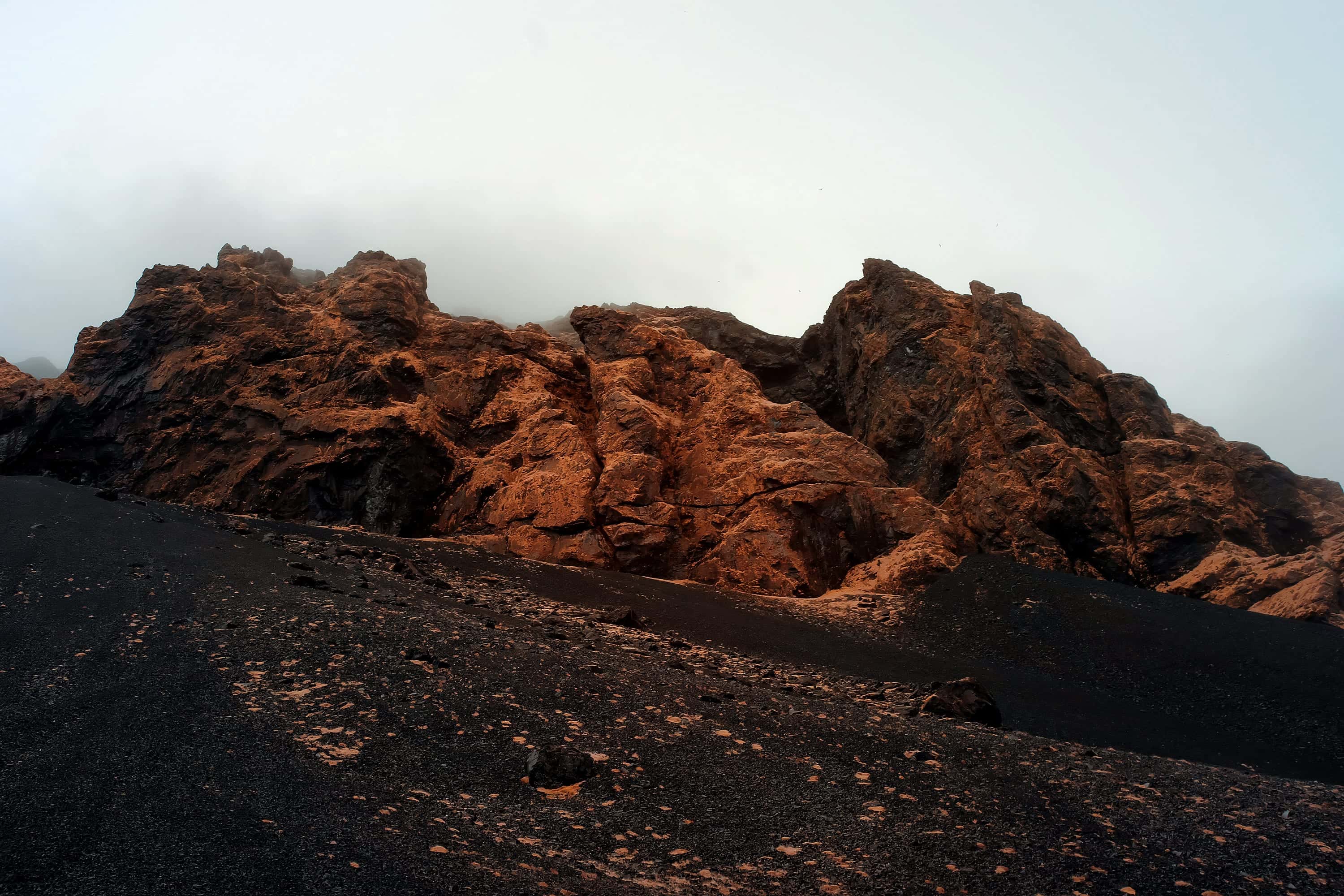 Rust-orange rock outcrops above a black ash slope dissolving into fog